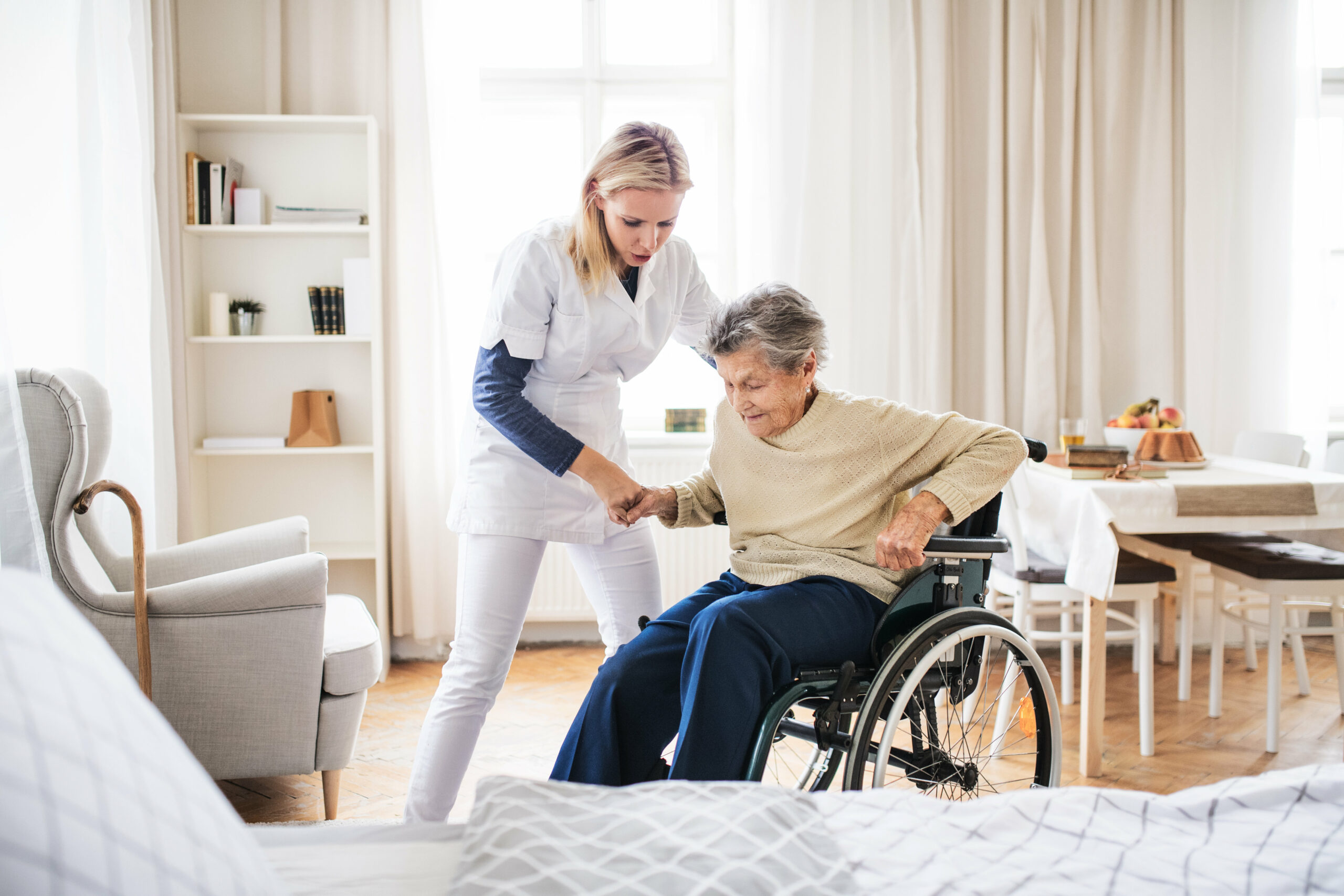 A health visitor helping a senior woman to stand up from a wheelchair.